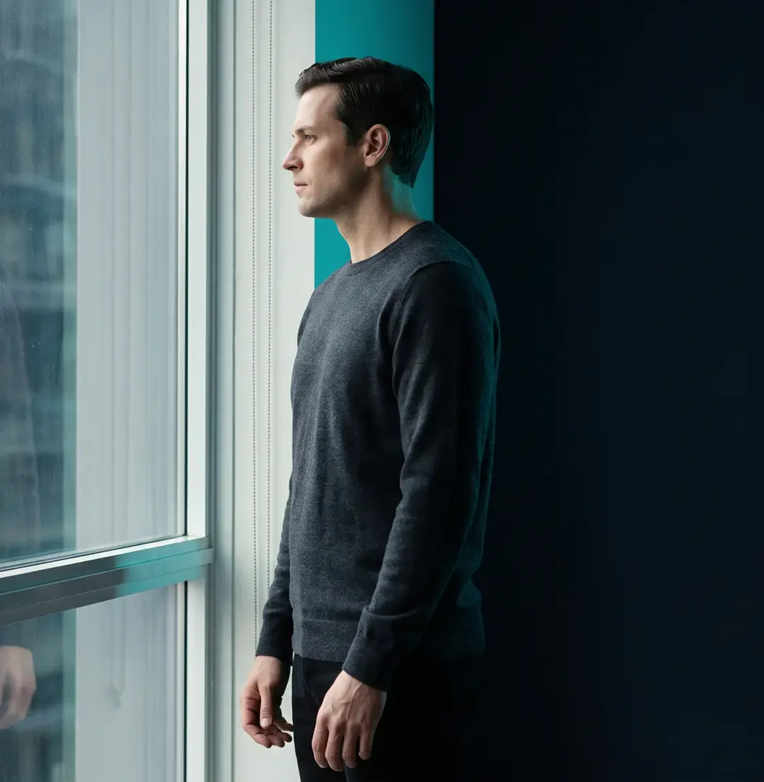 A man standing at a tall clinic window in profile, morning light on his face.