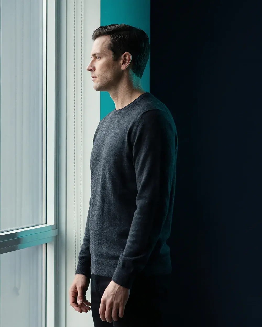 A man in his forties standing in profile against a teal panel, cool rim light along his jaw.